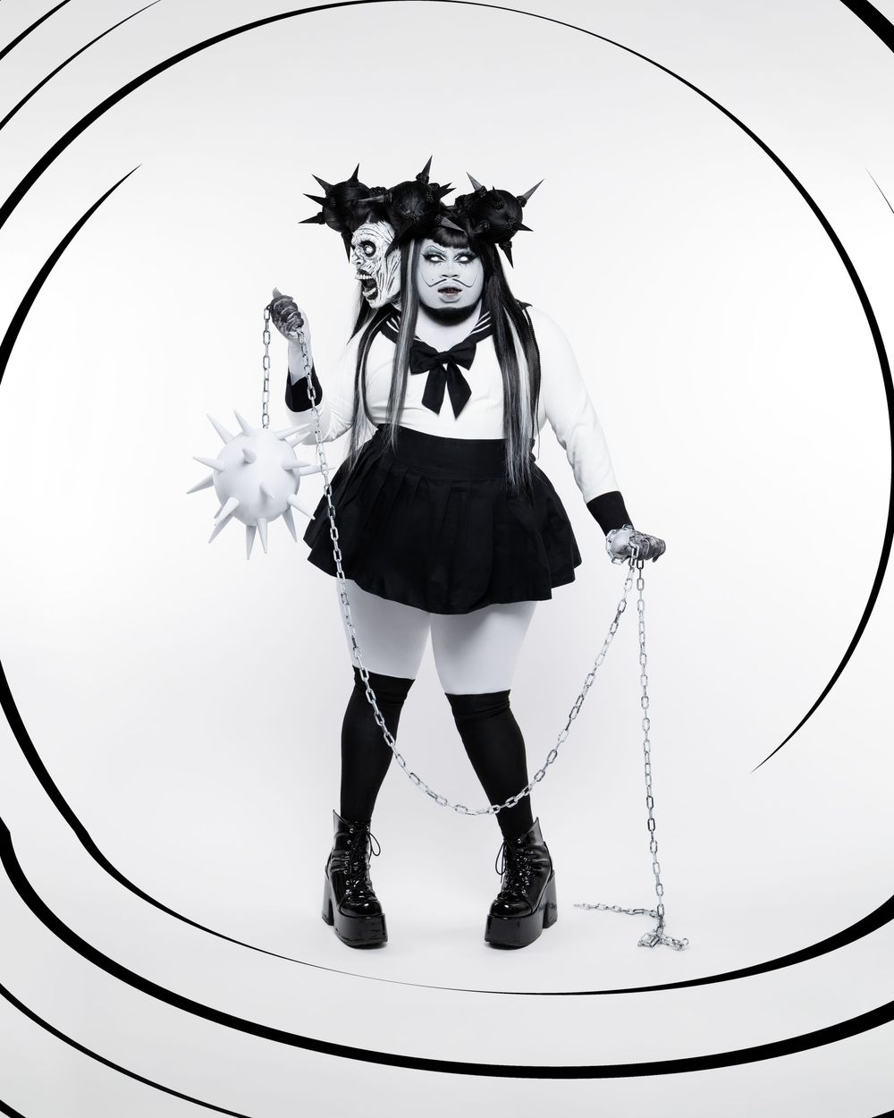 Gothic cosplay model in black dress and boots poses with spiked props against white circular background.
