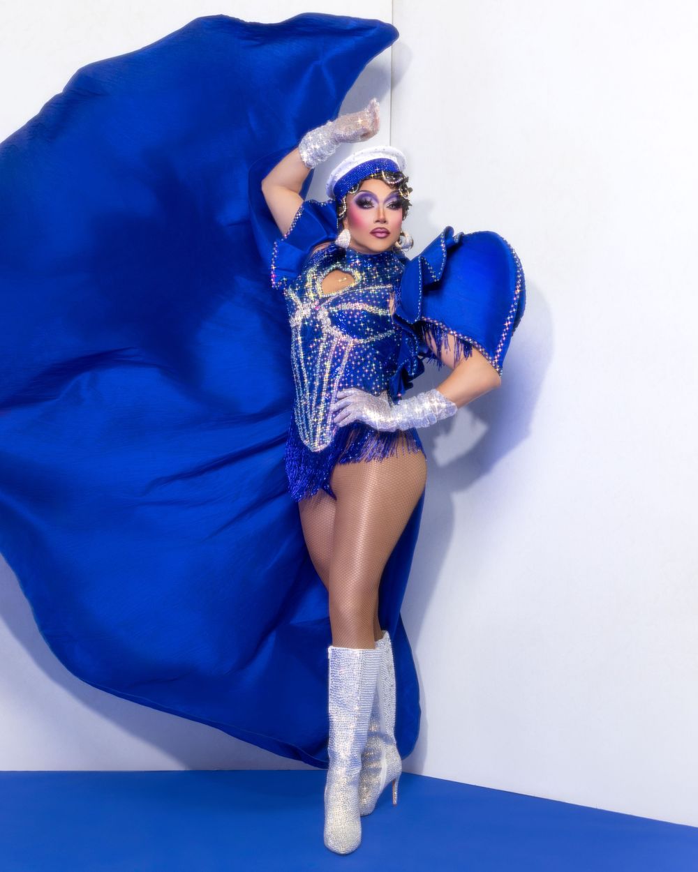 Performer in a dazzling blue sequined bodysuit with dramatic flowing cape and white boots strikes a dynamic pose.