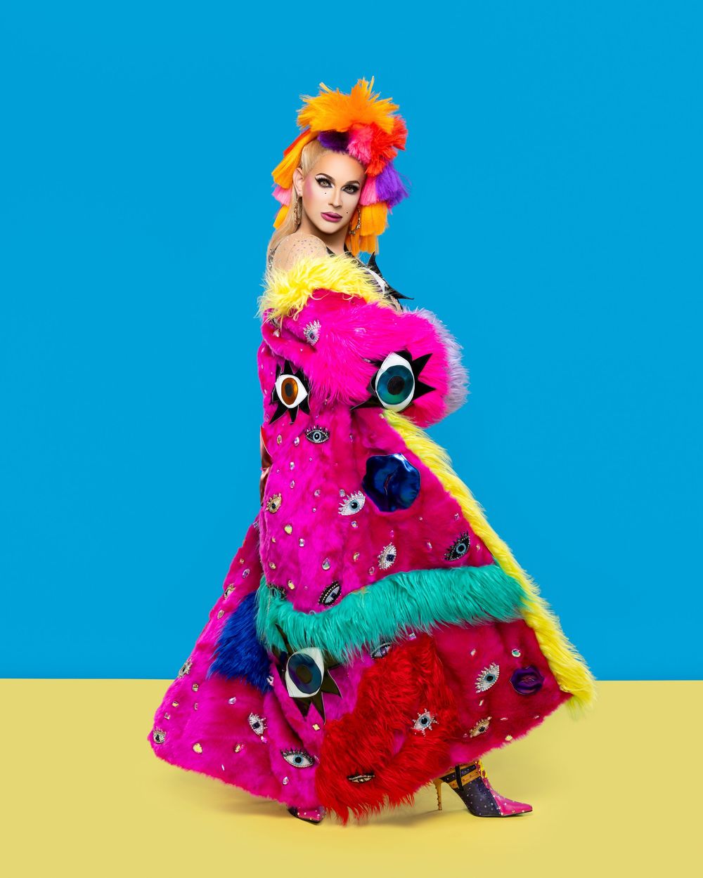 Artistic portrait against blue and yellow background featuring vibrant pink feathers and bold floral headpiece.