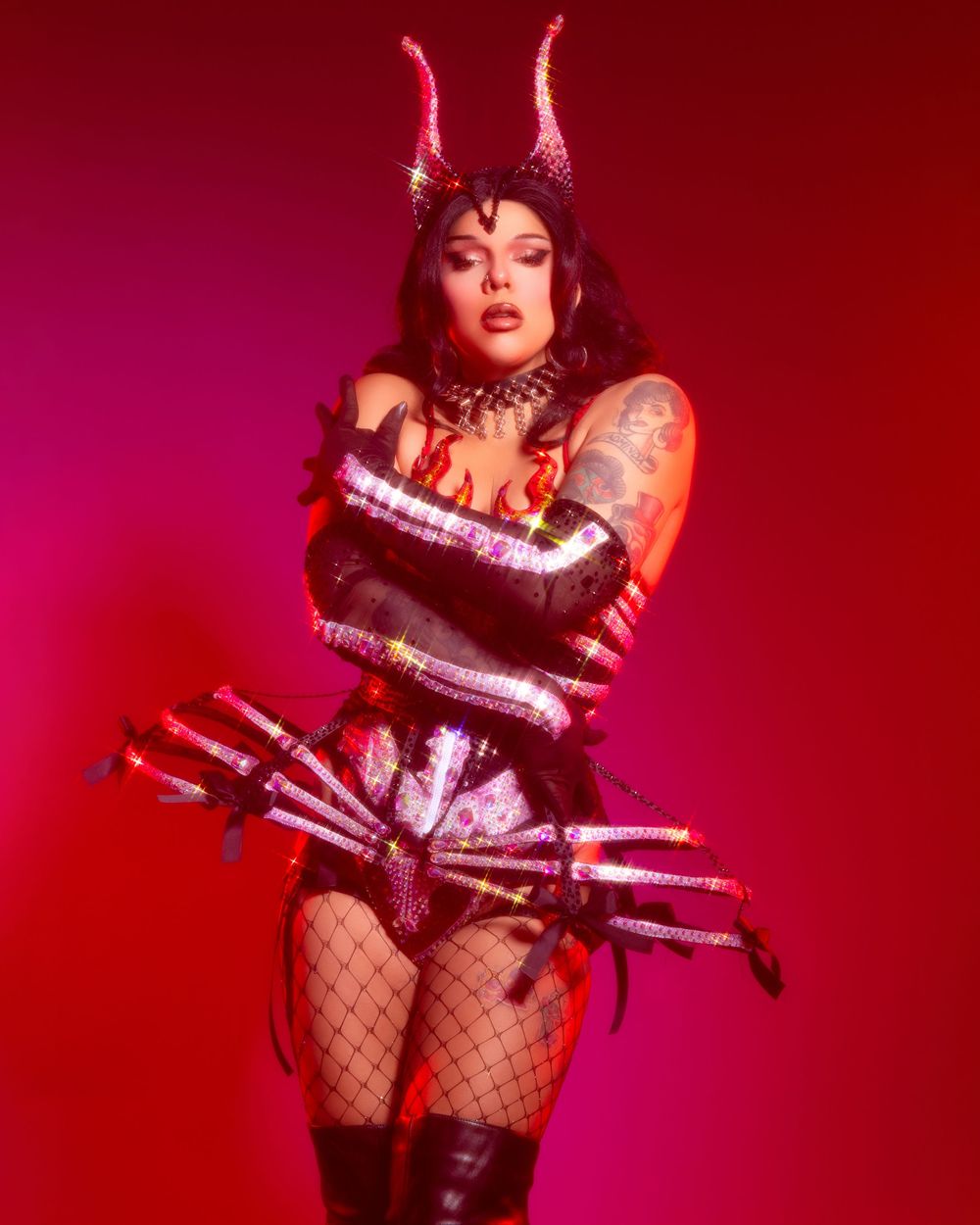 A performer in fishnets and dark costume poses dramatically against a red backdrop in a burlesque-style photoshoot.
