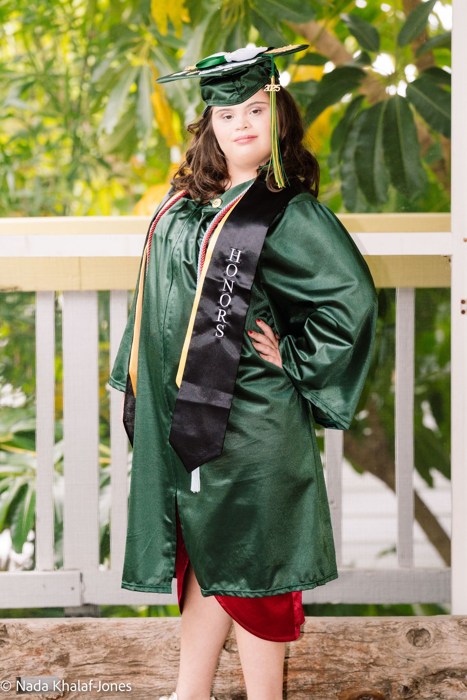 Graduate in dark green cap and gown poses outdoors near white railing and tropical foliage.