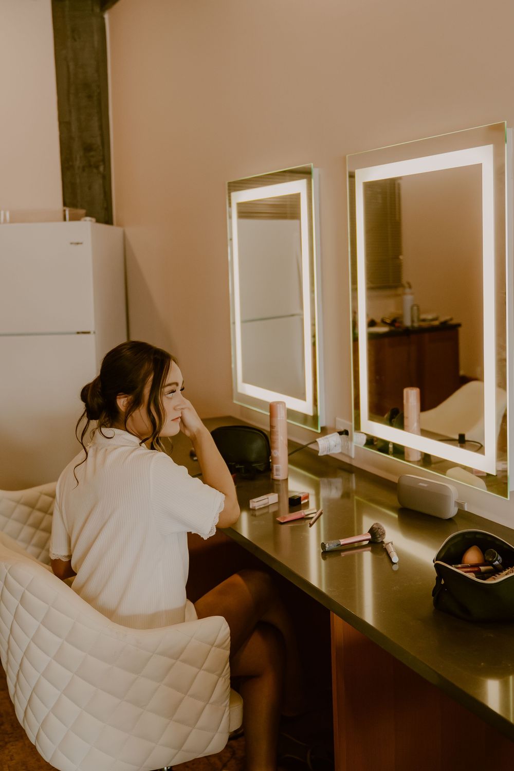 A person in a white quilted jacket sits at a mirrored vanity station in what appears to be a salon or makeup area.