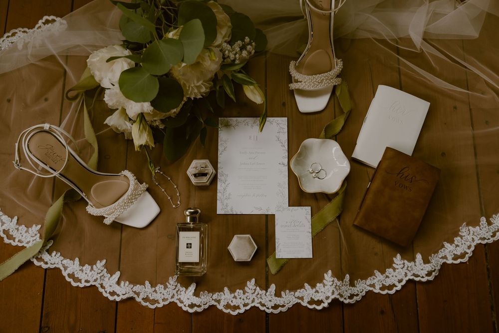 Wedding details flat lay with lace veil, stationery, flowers and accessories arranged on wooden surface.