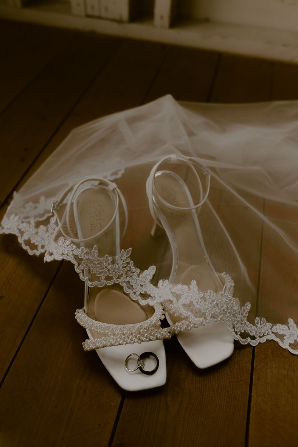 A delicate white bridal veil trails behind elegant white wedding shoes on a dark wooden floor.