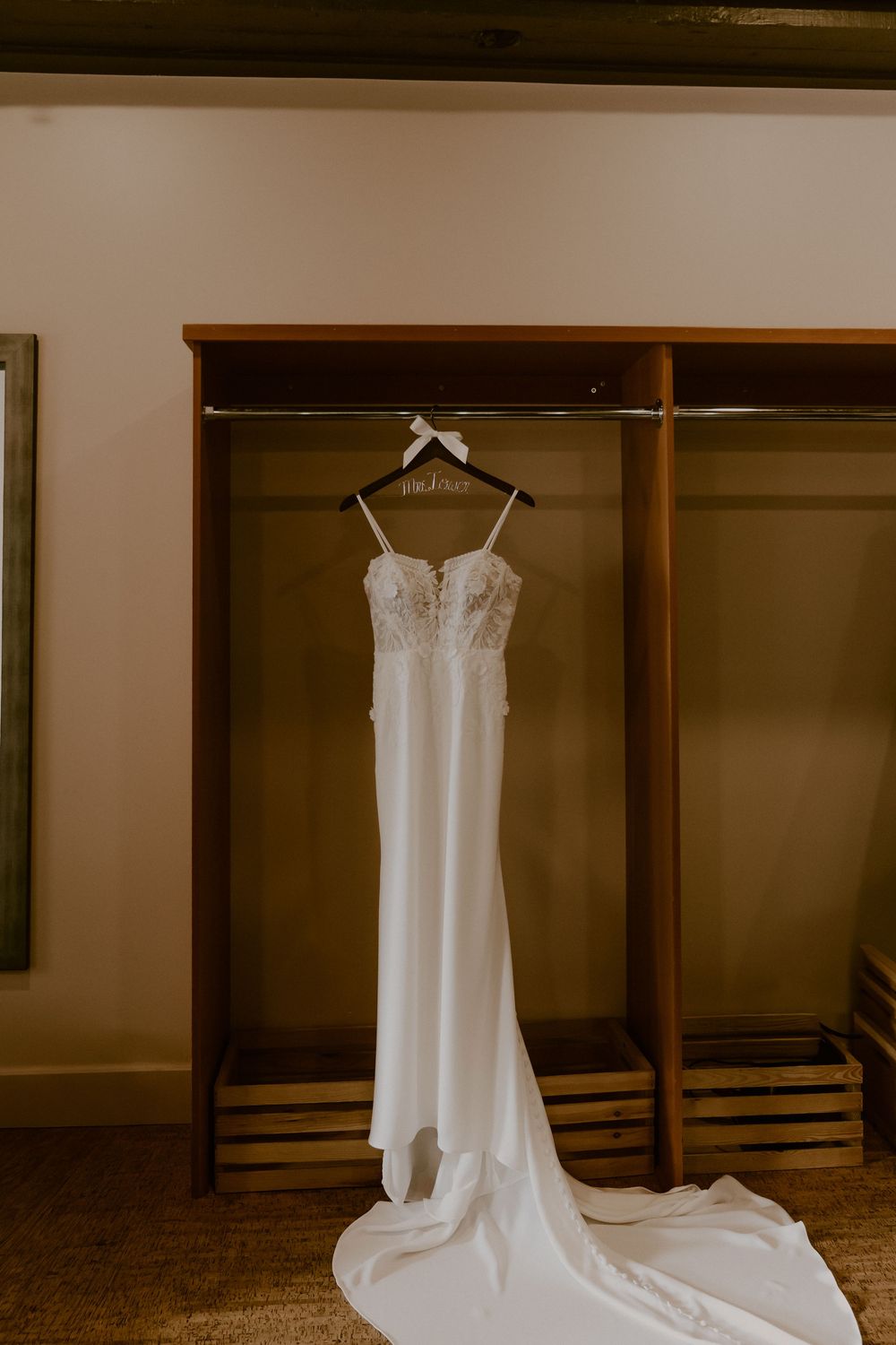 Elegant white wedding dress with lace bodice and flowing train hanging in wooden wardrobe against neutral wall.