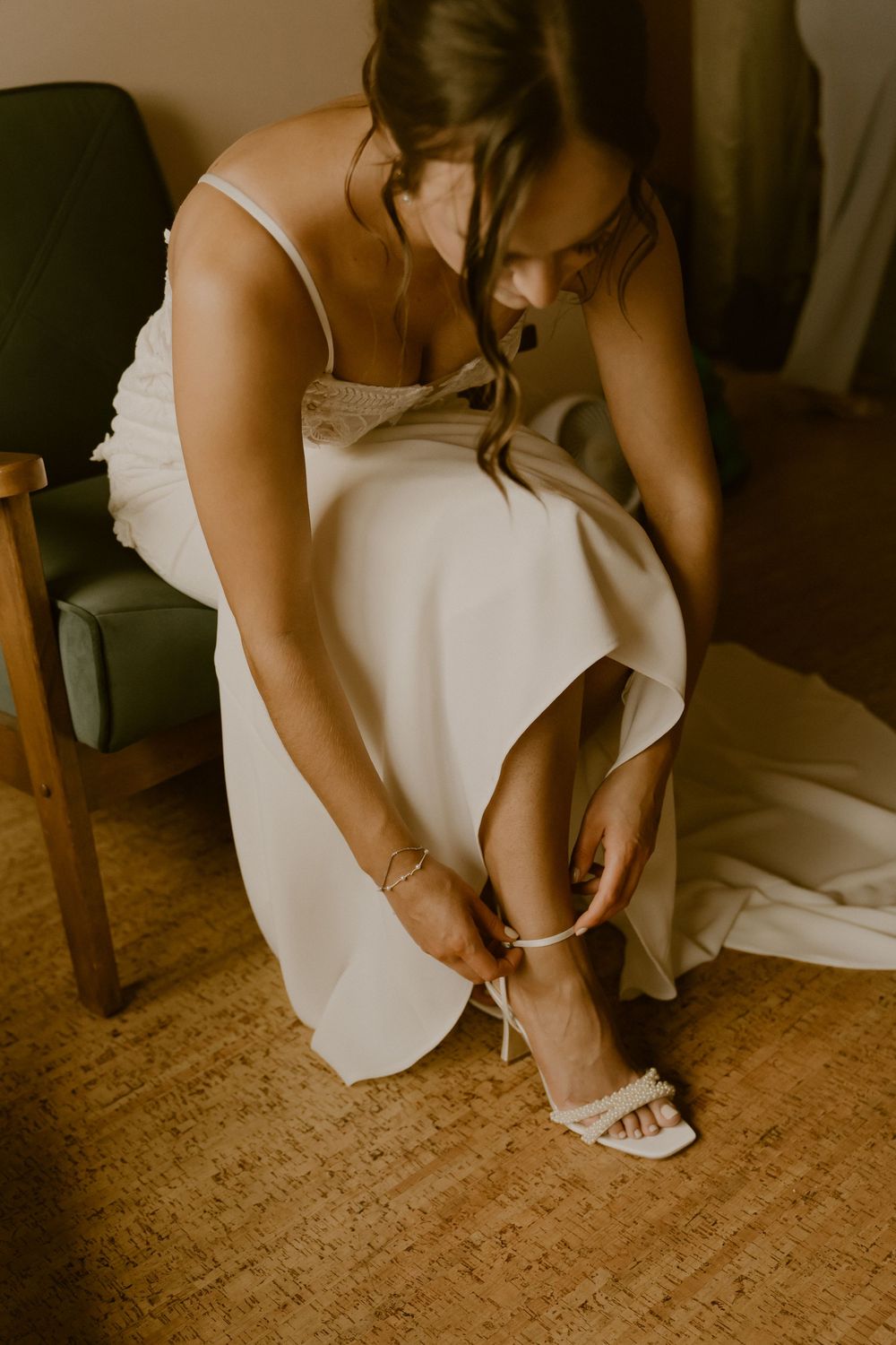 A person in a white dress fastens strappy sandals while sitting in a warmly lit room.