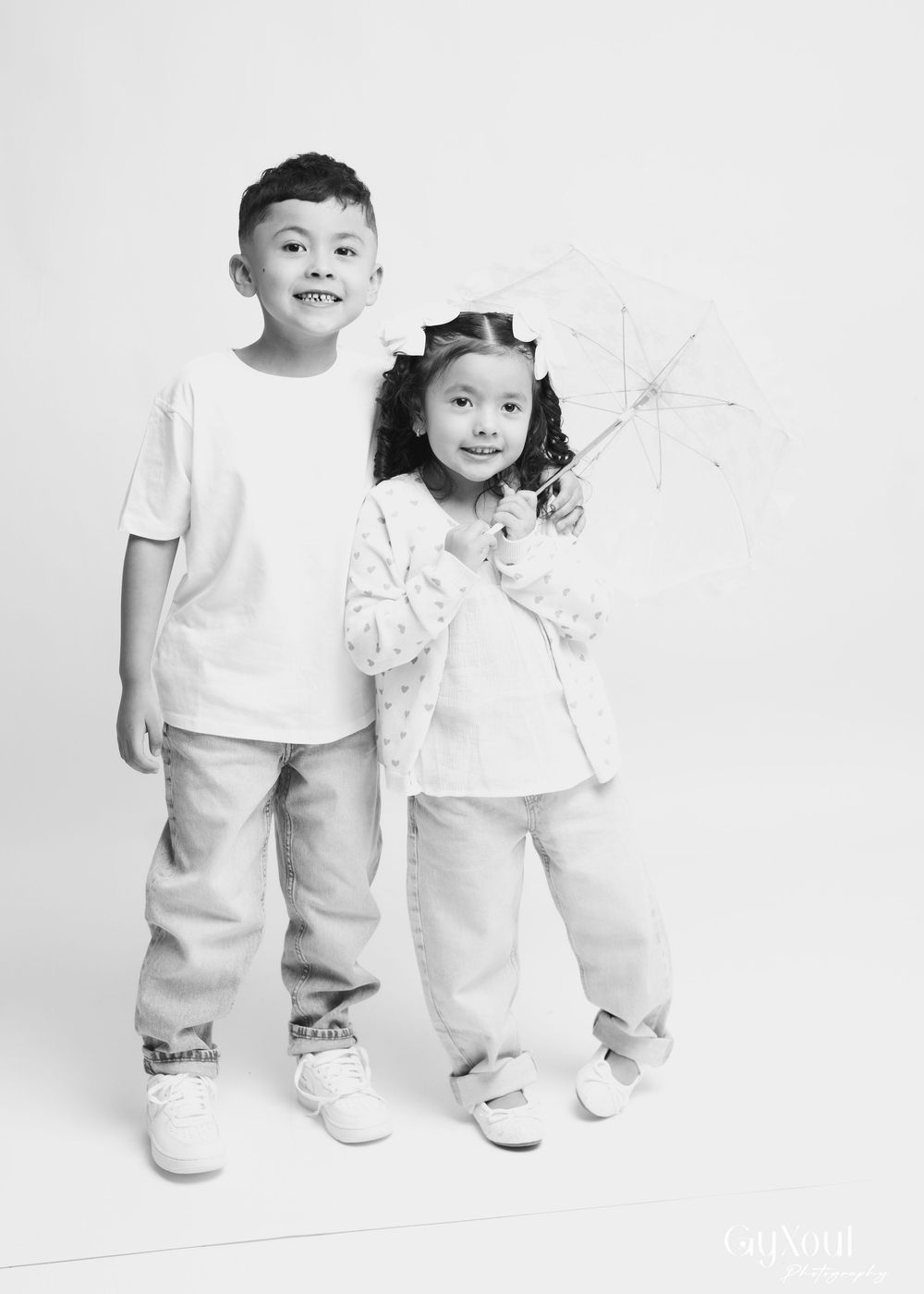 Three children in white outfits stand together against a white background in a minimalist studio setting.