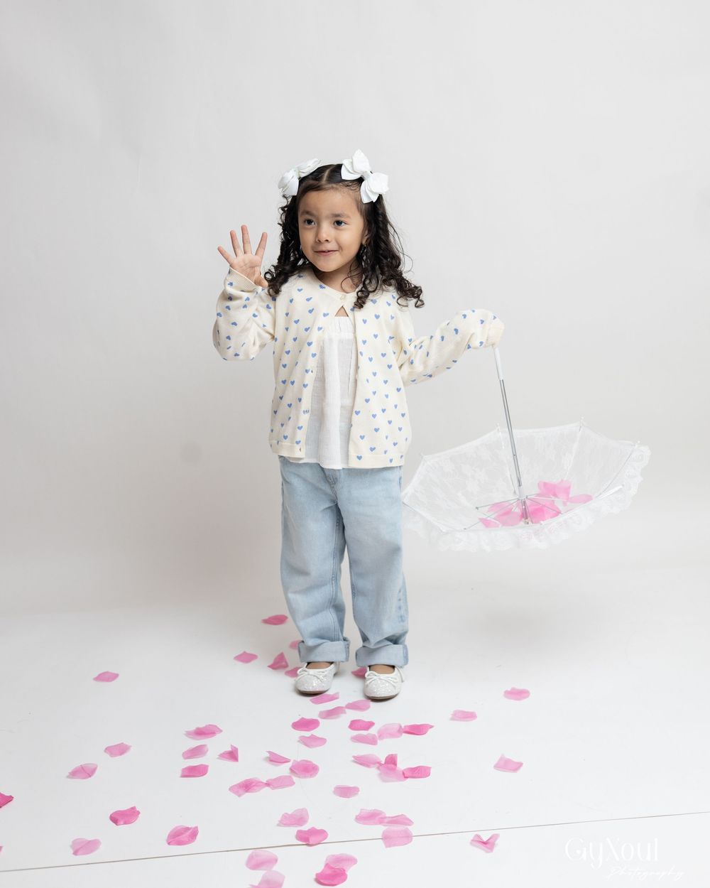 A small child wearing a floral cardigan and jeans plays with pink paper hearts scattered on a white floor.