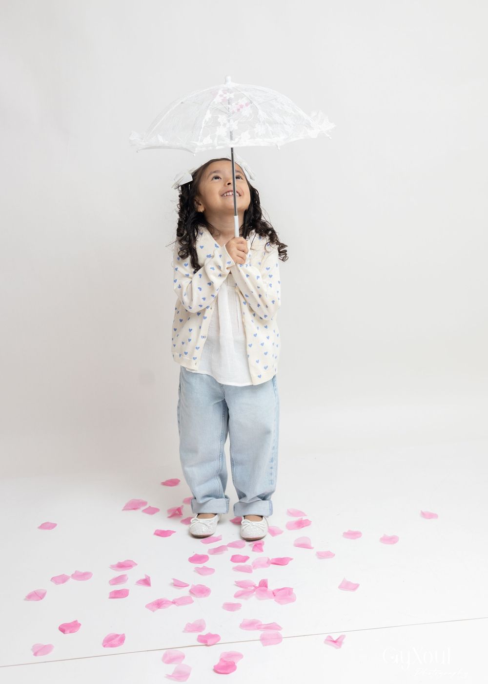A person in a white polka dot shirt and jeans dances playfully on a white floor scattered with pink paper hearts.