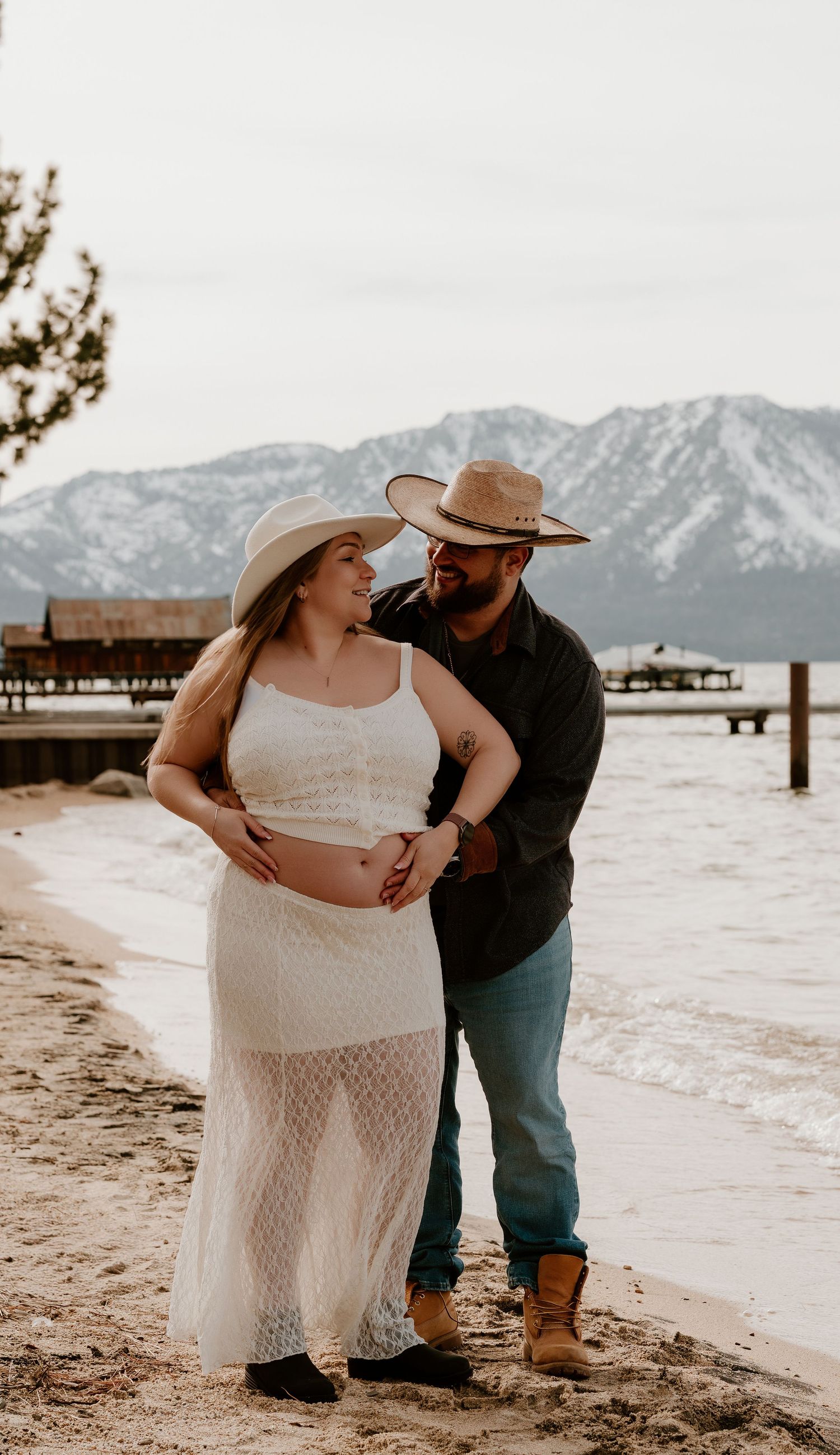 A couple embraces on a beach with snow-capped mountains in the background at Lake Tahoe.