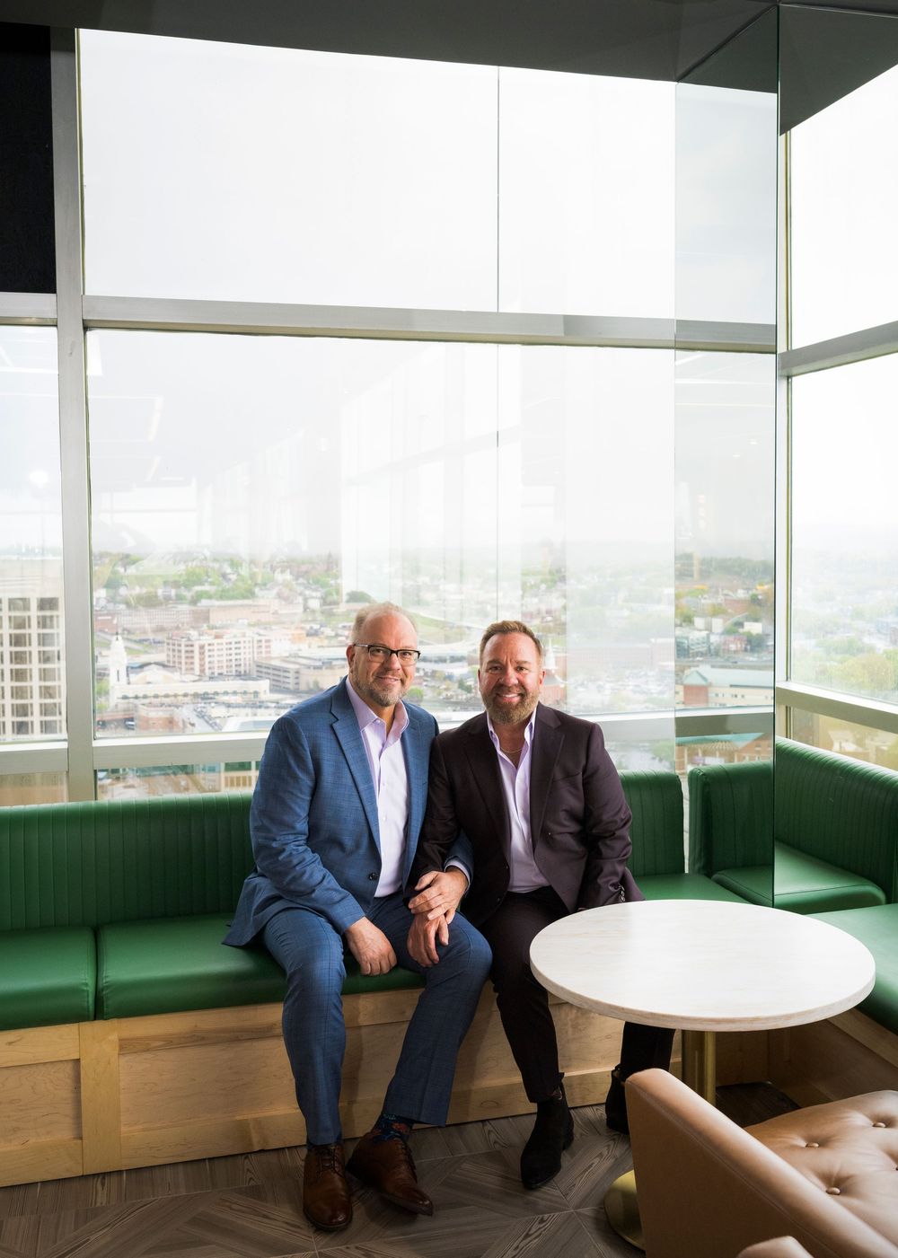 Two grooms sit together on a green booth by large windows with a city view behind them.