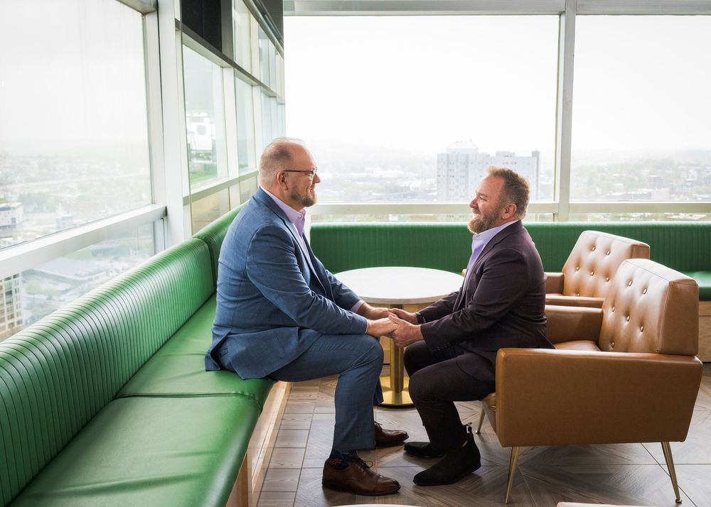 Two businessmen in suits having a professional meeting on a green booth in a modern high-rise office lounge.