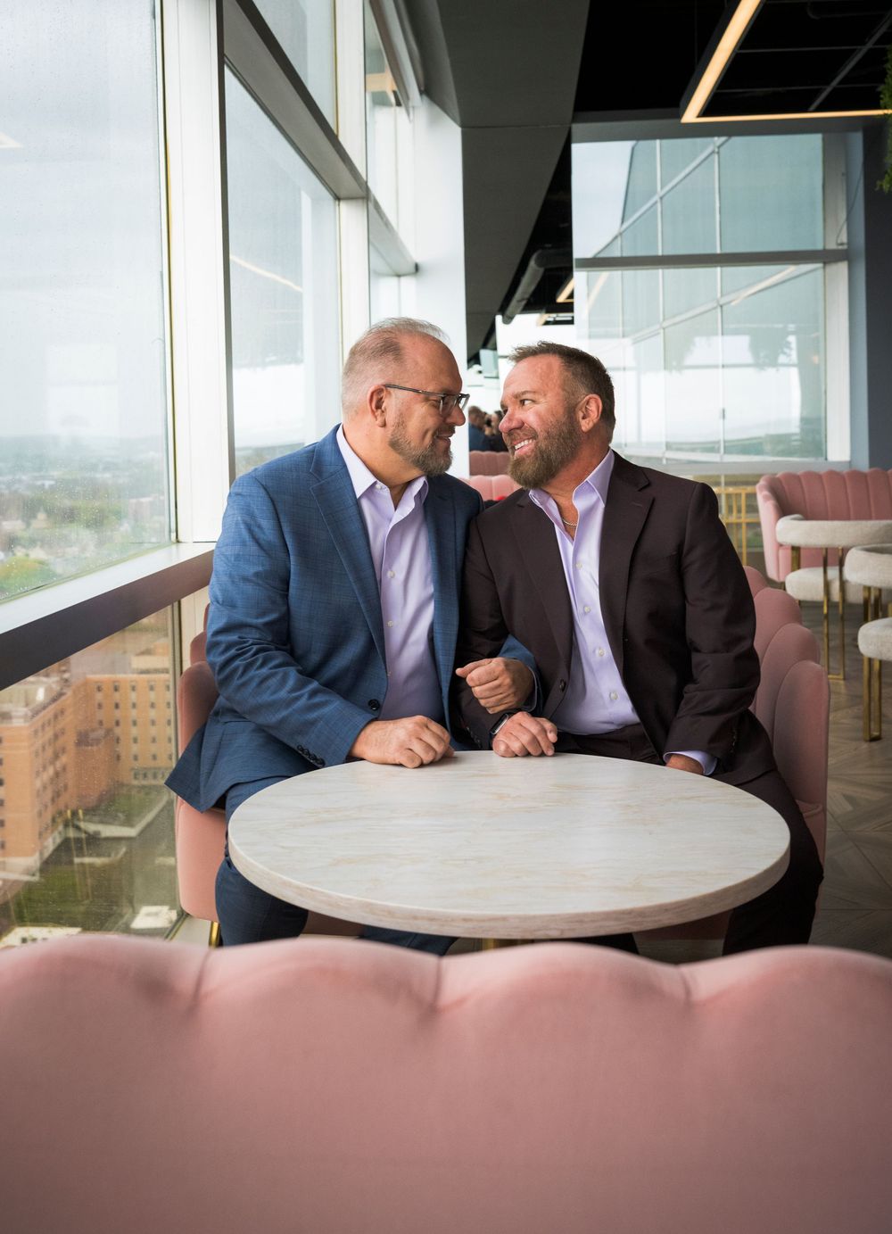 Two grooms share an intimate moment at a round marble table in a rooftop venue.