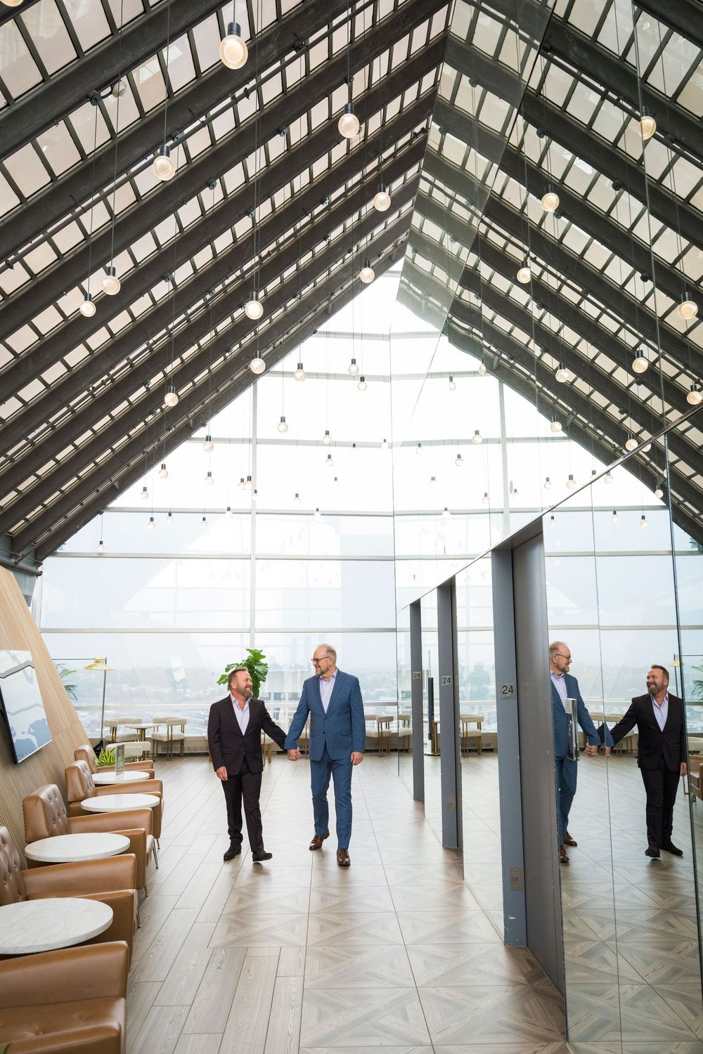Two grooms hold hands walking through an elegant glass-ceiling wedding venue with herringbone floors.