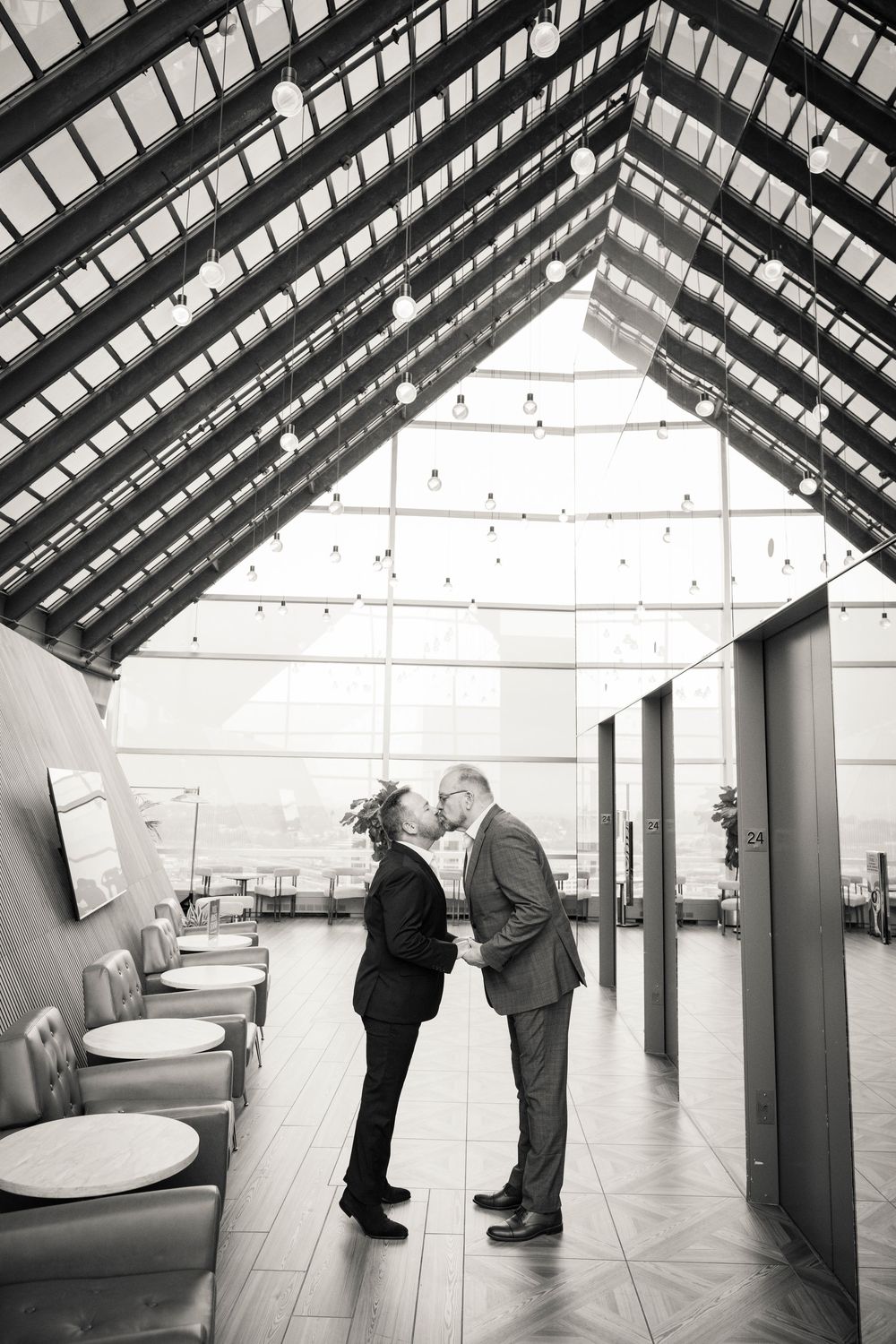 Two grooms share a romantic kiss in a modern venue with a glass ceiling and string lights.