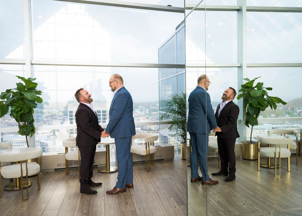 Two businessmen shaking hands in a modern high-rise office lounge with floor-to-ceiling windows and city views.