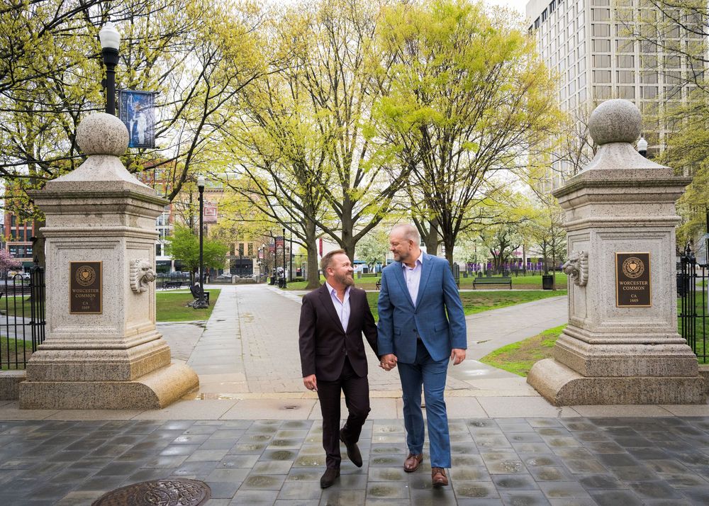 Two men holding hands walk through ornate stone gates in a park with spring trees.