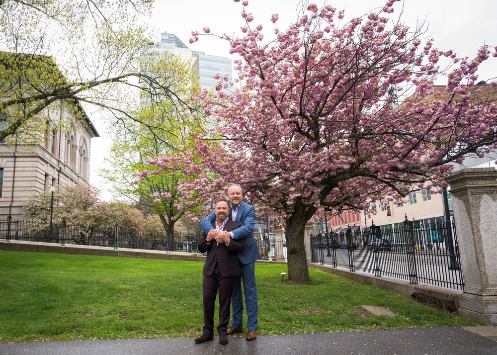 Two men pose together under blooming pink cherry blossom trees in a park setting with iron fence and buildings behind.