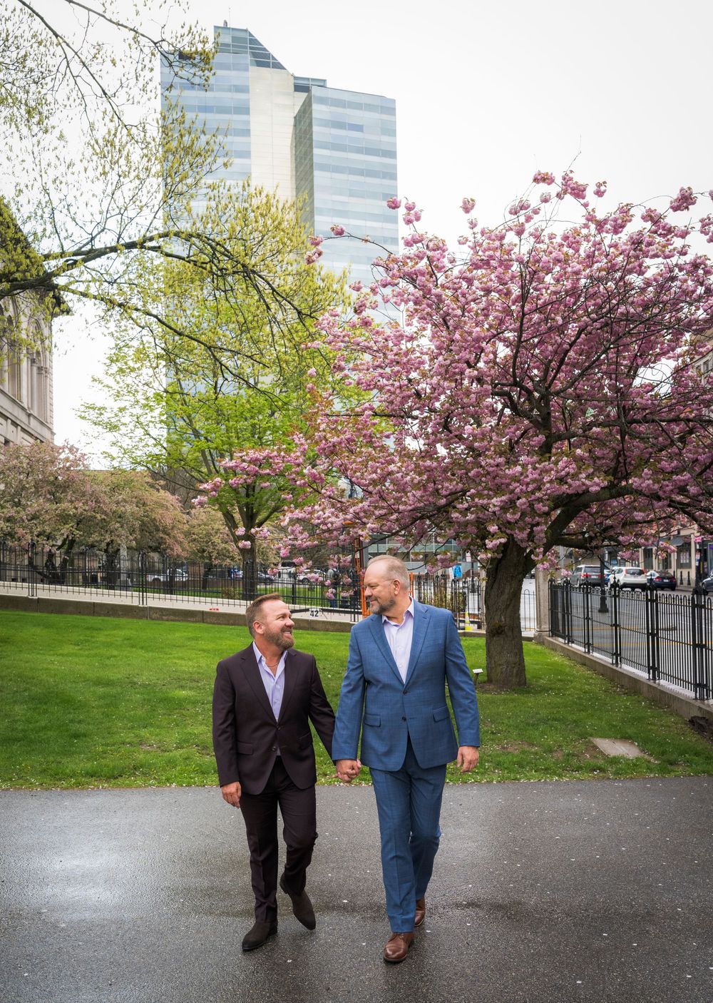 Two grooms walk hand in hand past blooming cherry blossom trees in a city park.
