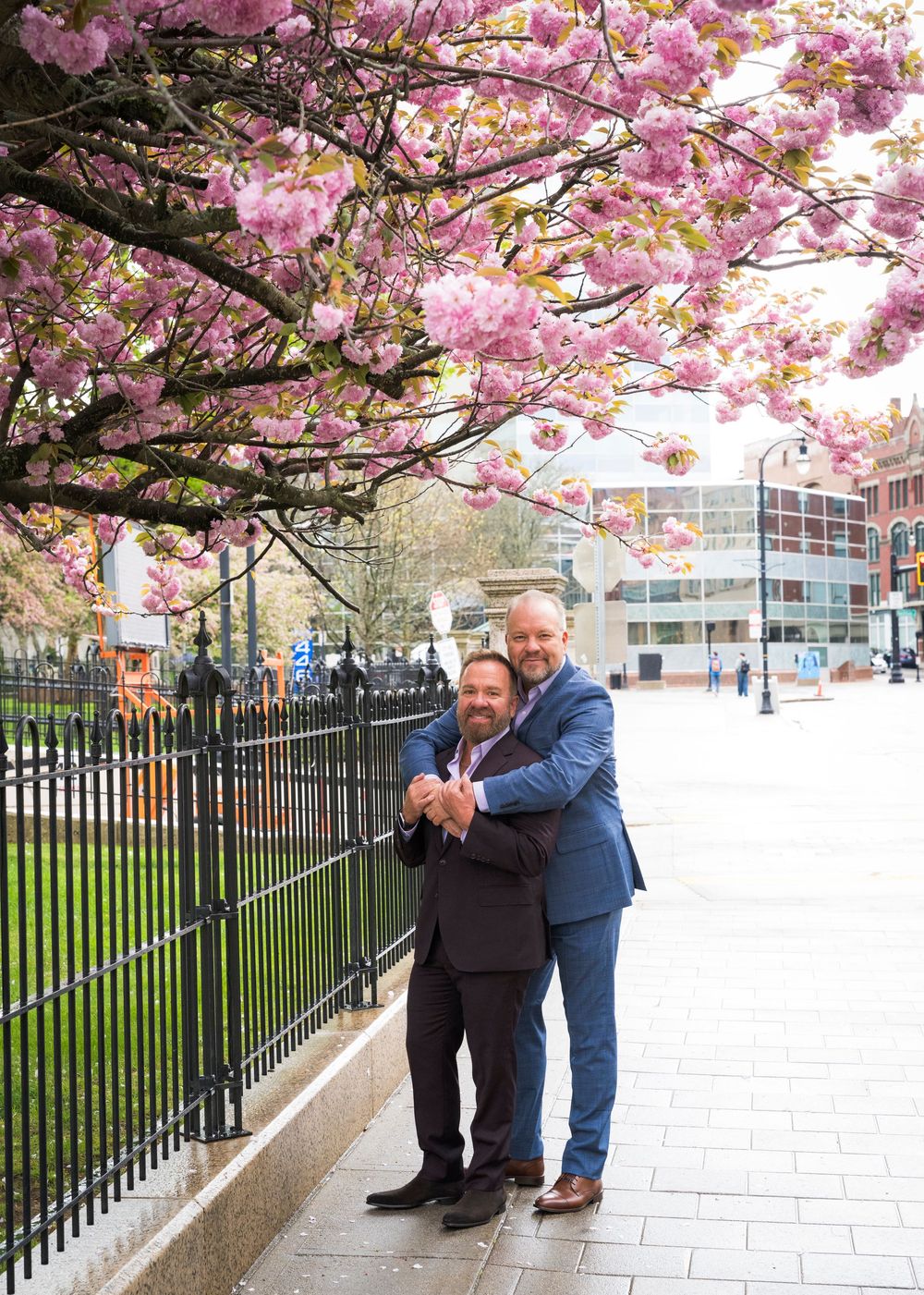 Two grooms embrace under pink cherry blossoms beside iron fence in spring outdoor wedding photo.