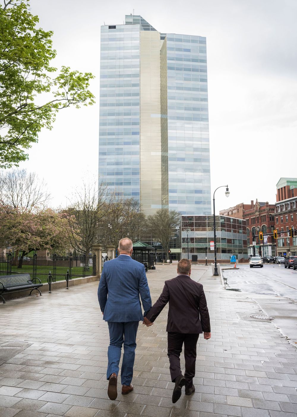 Two men holding hands walking toward a modern glass skyscraper in an urban plaza.