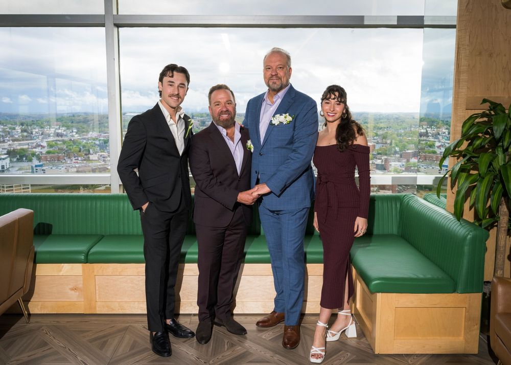 Two grooms pose with family members in front of panoramic city view windows at wedding reception.