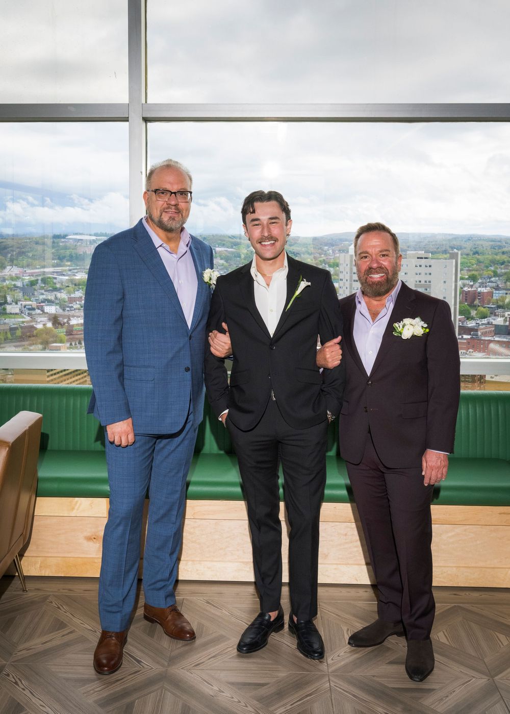 Three men in suits with boutonnieres pose together at a rooftop venue with city views.
