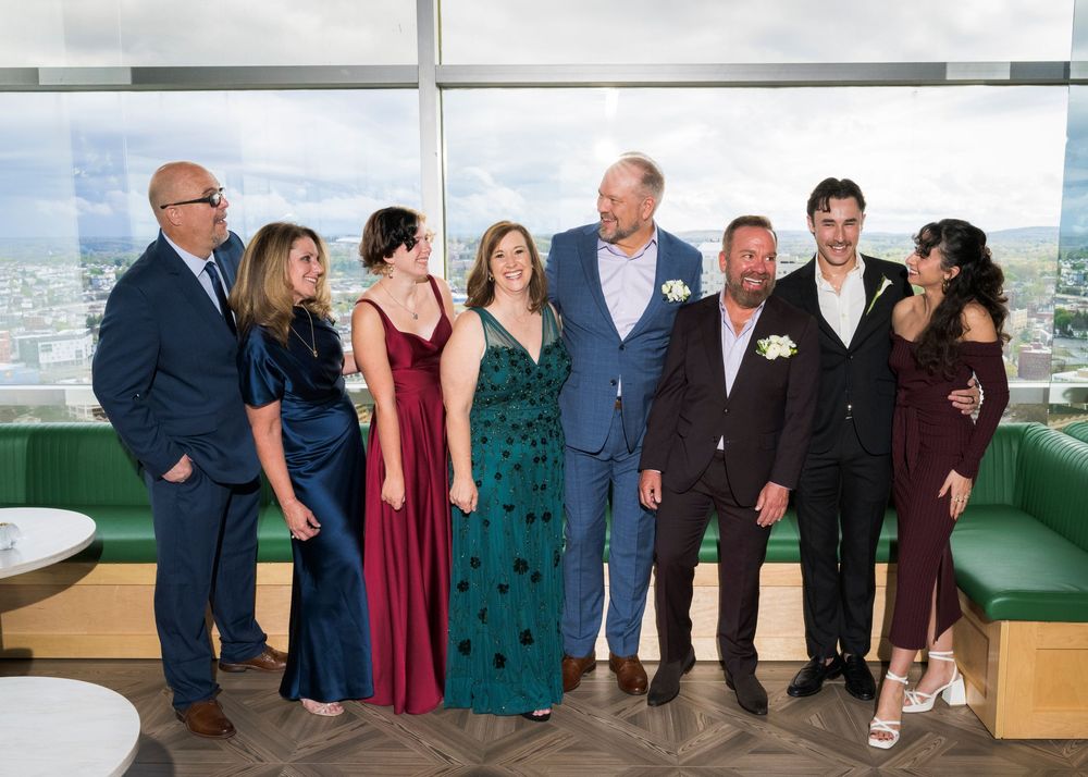 Group of eight elegantly dressed wedding guests pose together at a rooftop reception venue.