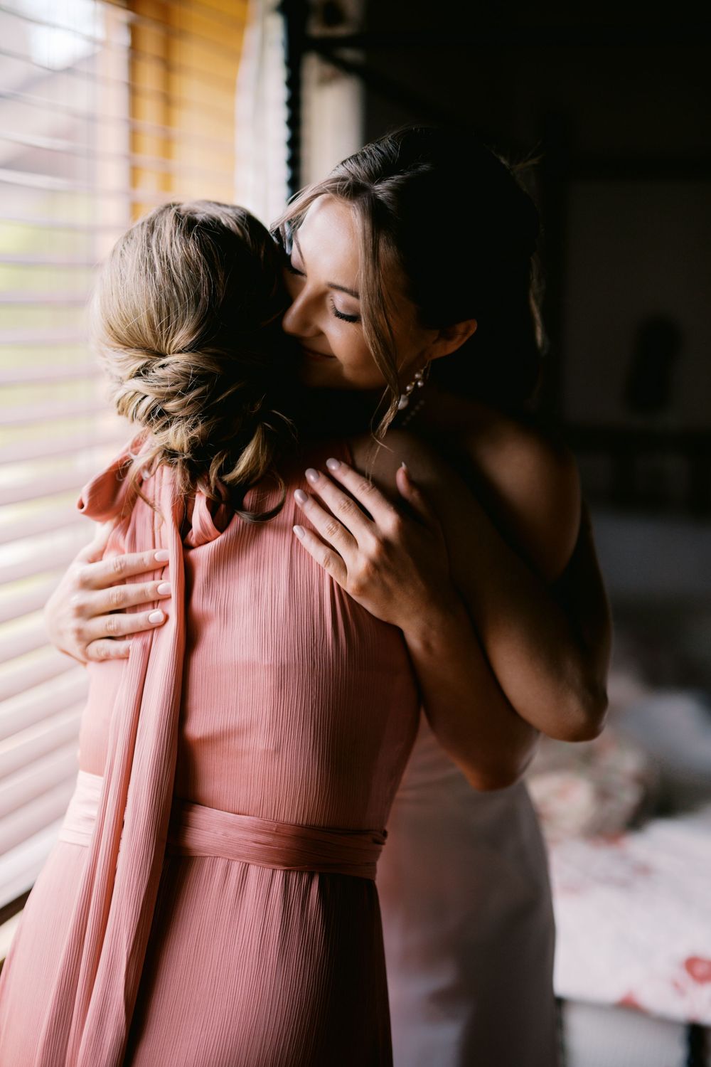 Two people share an intimate embrace while wearing a pink dress in a softly lit room by a window.