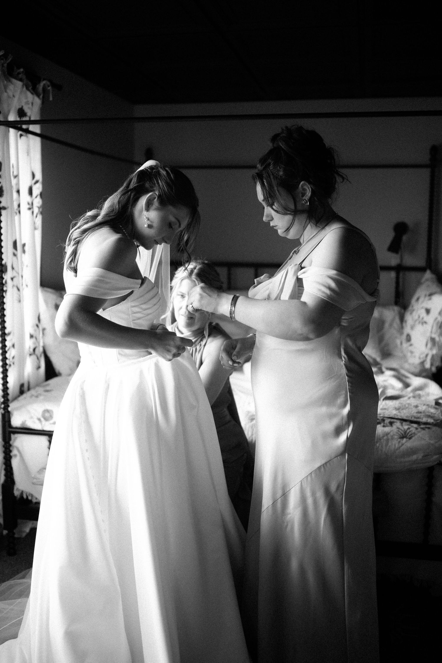Black and white photo showing bridesmaids helping each other get ready in white dresses.