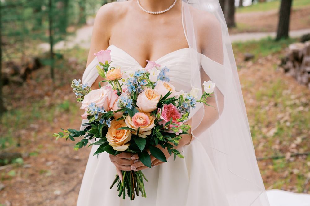 A stunning bridal bouquet of peach roses and greenery is held against a white wedding dress in an outdoor forest setting.