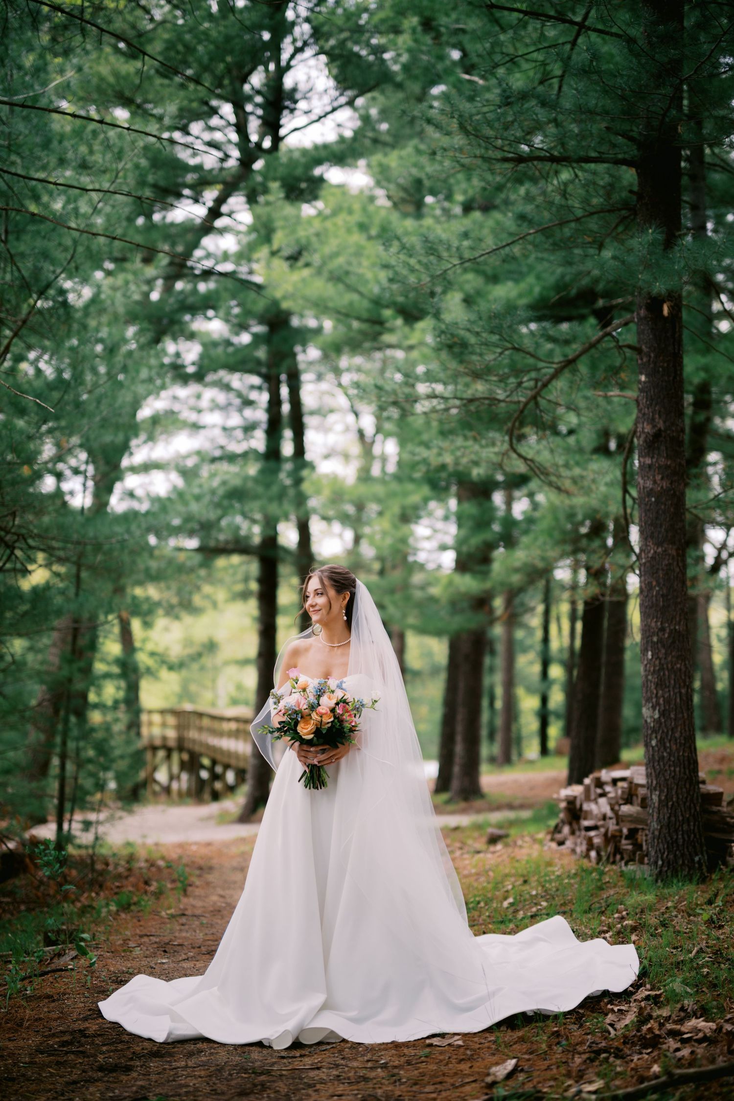 A flowing white wedding dress and autumn bouquet create a romantic scene along a pine tree-lined forest path.