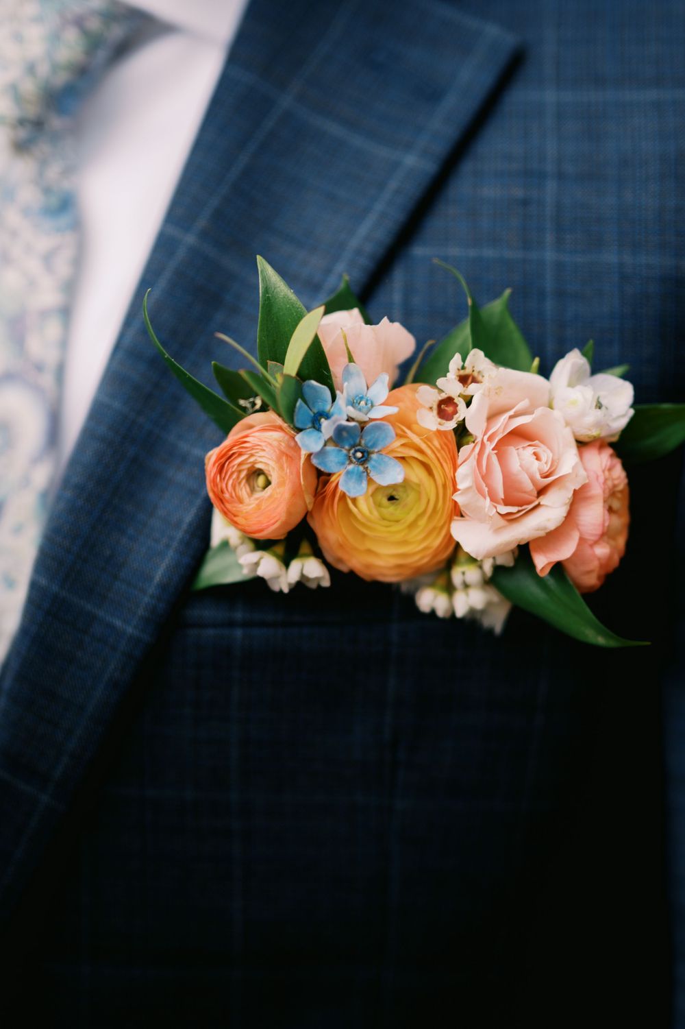 A close-up of a boutonniere featuring peach ranunculus, blue delphinium, and baby's breath against a navy suit jacket.
