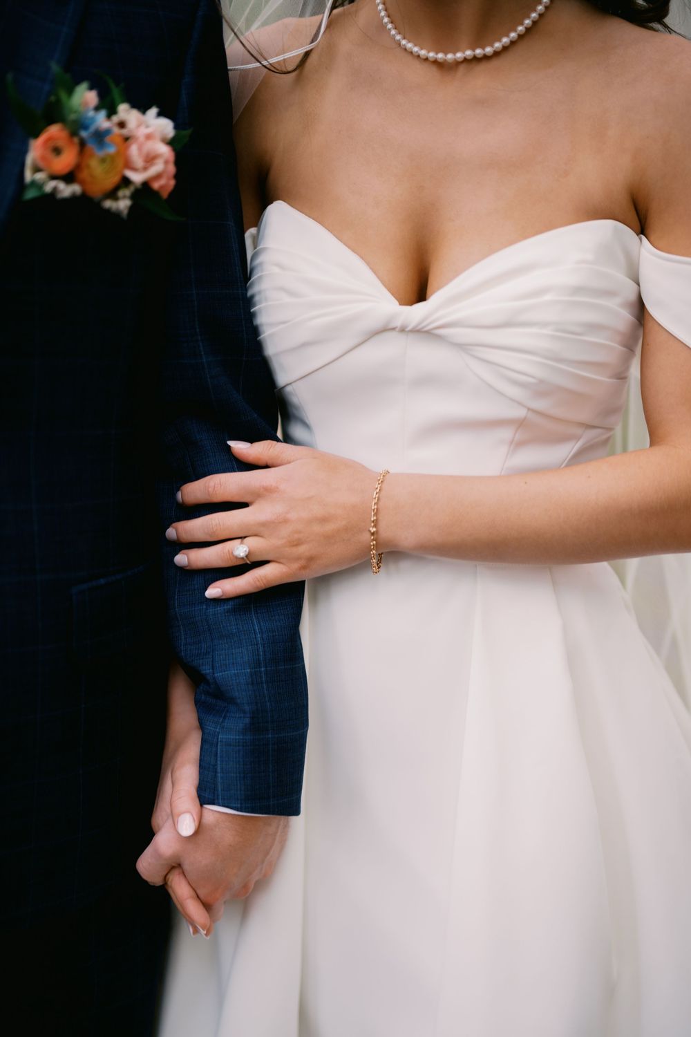 Close-up details of a white wedding dress and pearl necklace with embracing couple showing elegant bridal styling.