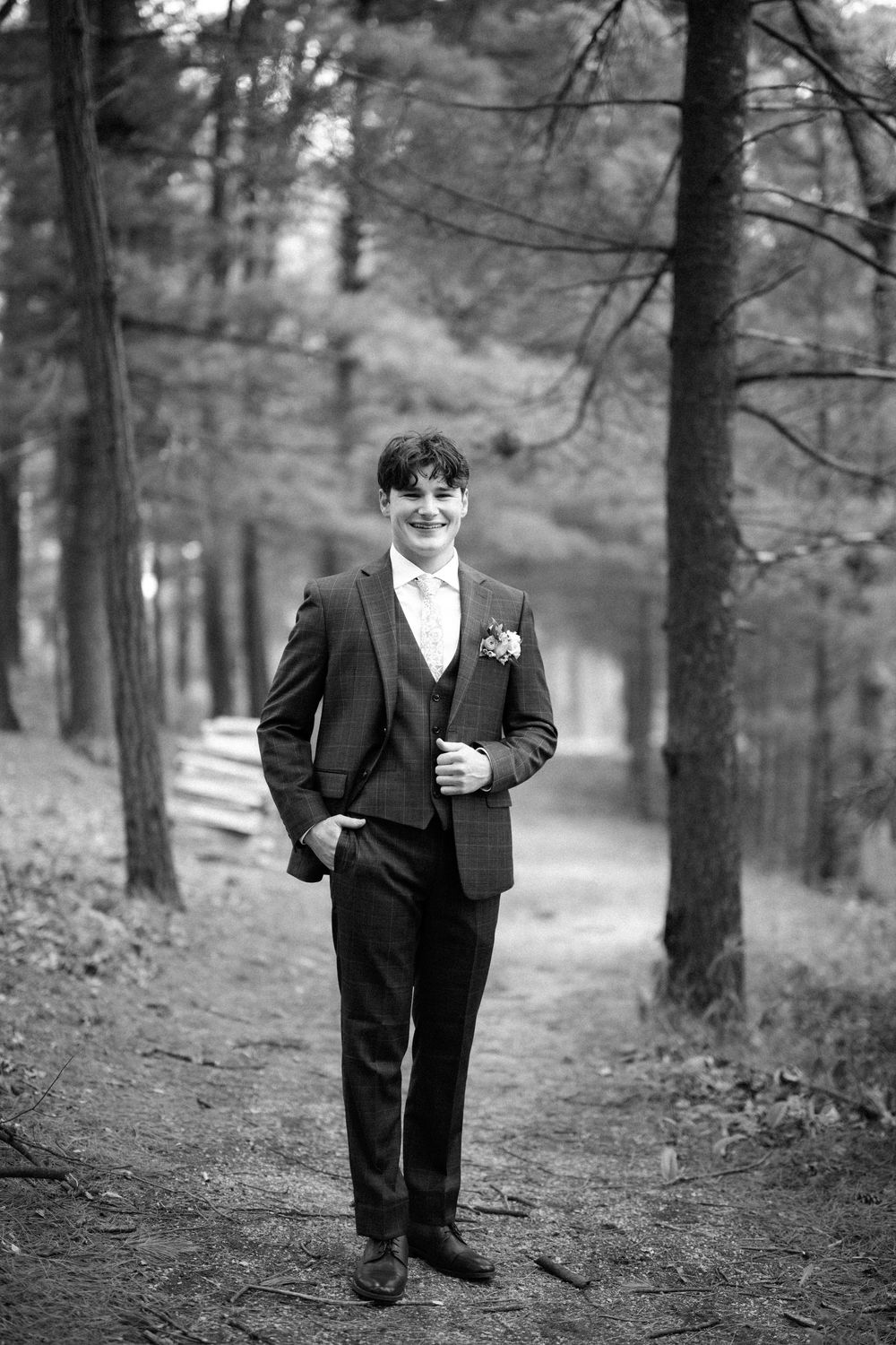 A groom in a classic black tuxedo stands smiling on a wooded path during a wedding photoshoot.