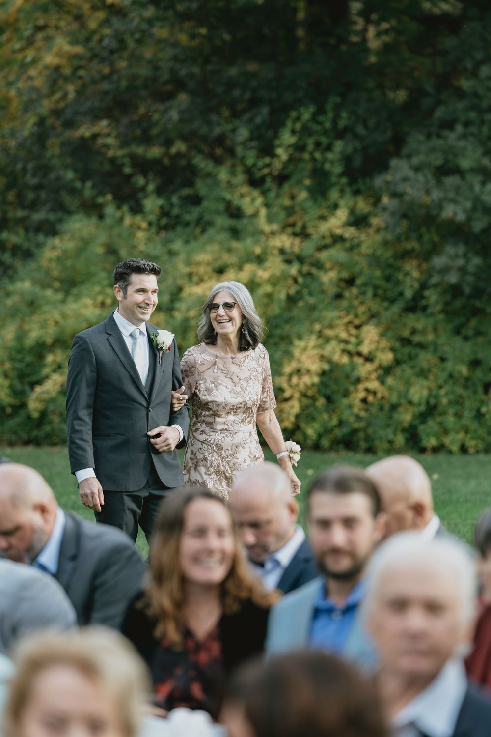 Wedding guests gather outdoors in a natural setting with autumn foliage in the background.