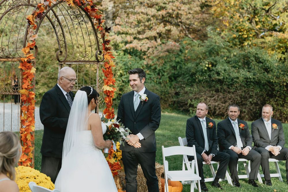 Outdoor fall wedding ceremony with orange floral arch and white chairs arranged on grass.