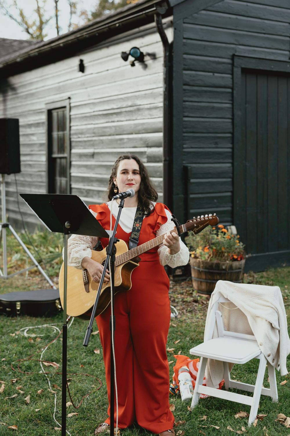 A musician in a red sweater performs with an acoustic guitar outdoors by a dark wooden building with white chairs.