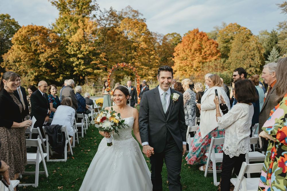 A fall wedding ceremony takes place outdoors with colorful autumn trees and white chairs arranged on a lawn.
