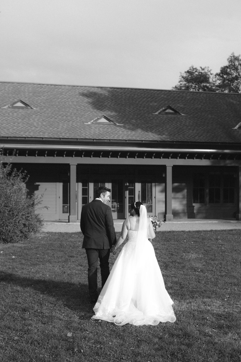 Black and white wedding photo sequence showing a couple walking across a grassy lawn in front of a long building.