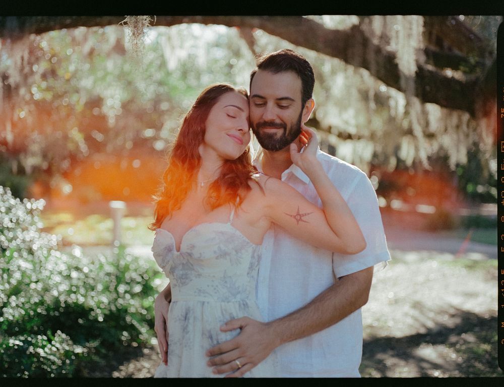 A couple embraces tenderly in a romantic outdoor setting with sunlit trees and foliage in the background.