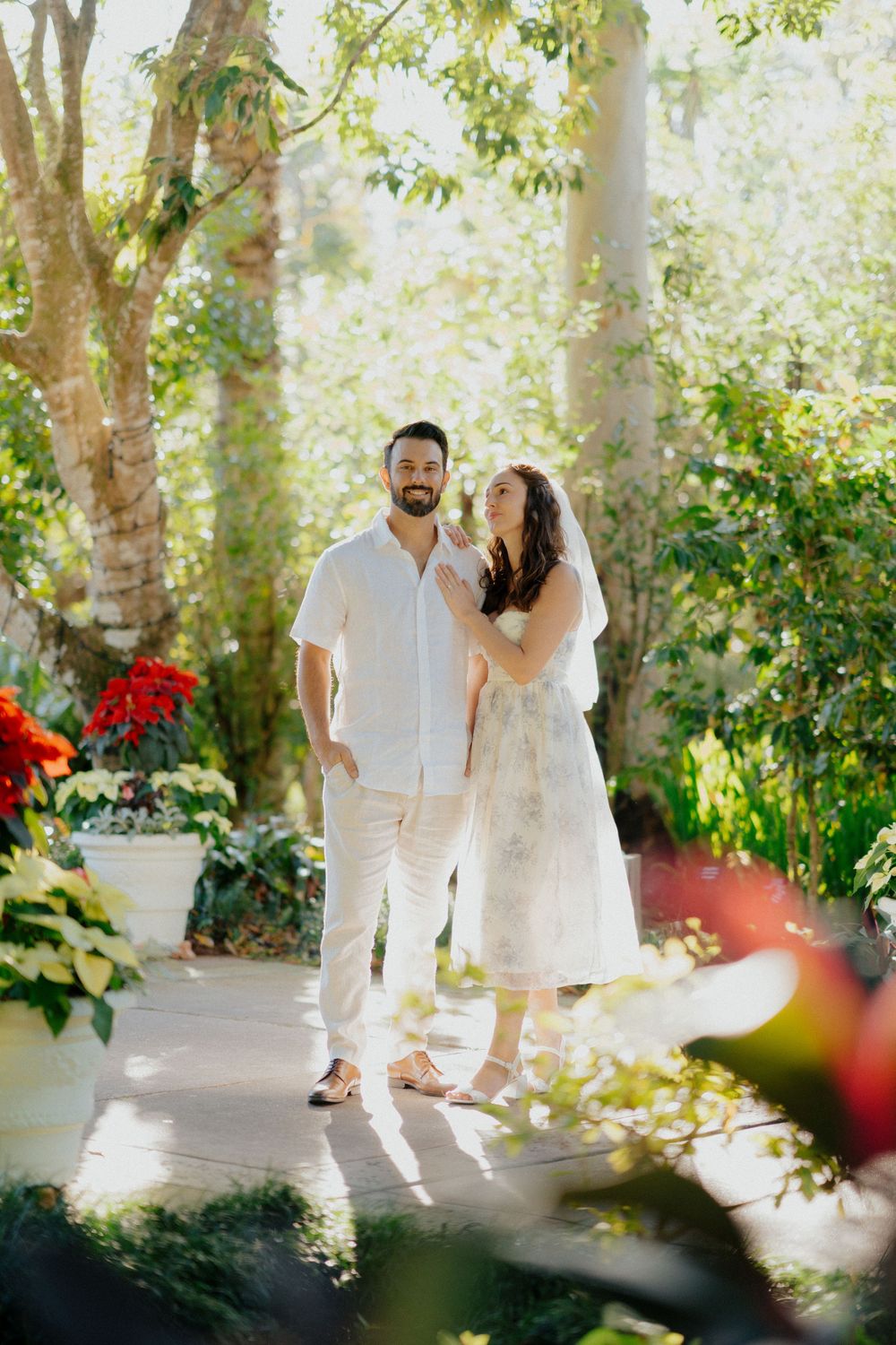A couple sharing a joyful moment together in a lush garden setting with red flowers and dappled sunlight.
