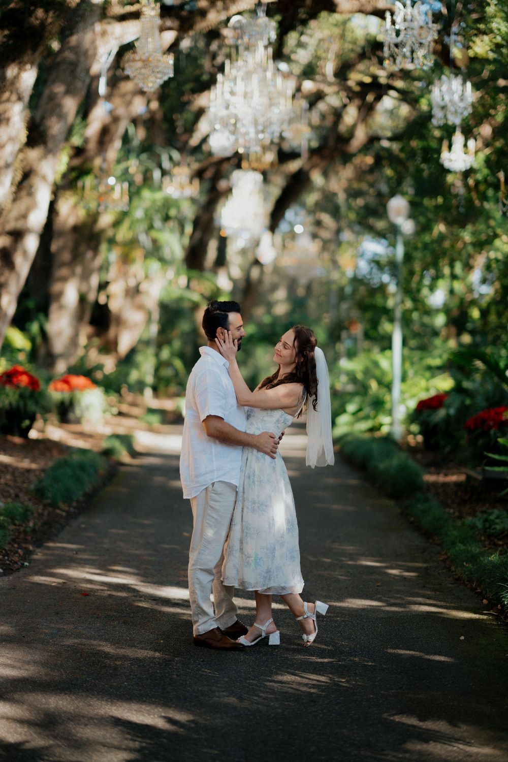 A couple in white attire dances together on a tree-lined path during a romantic outdoor photoshoot.