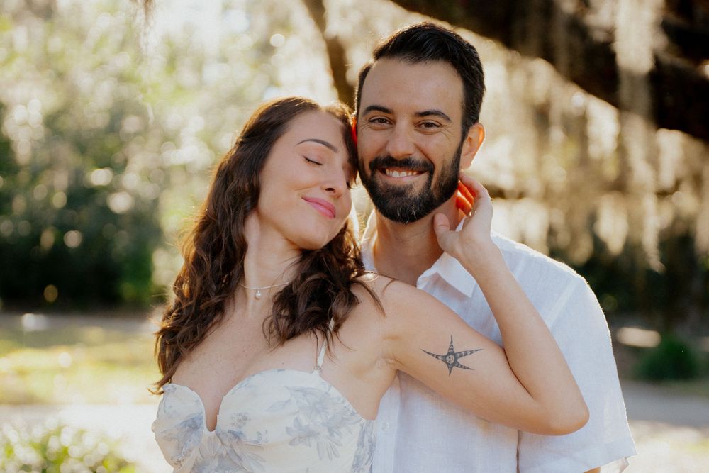 A romantic couple embraces outdoors in a sunlit garden setting wearing white clothing.