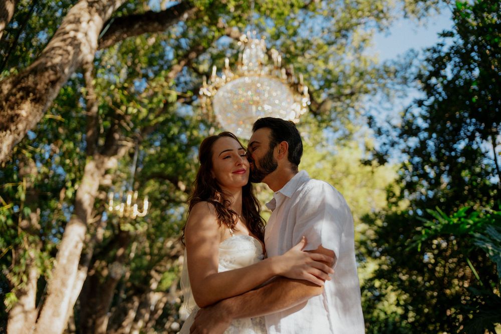 A couple embraces outdoors in a romantic moment surrounded by sunlit trees and greenery.