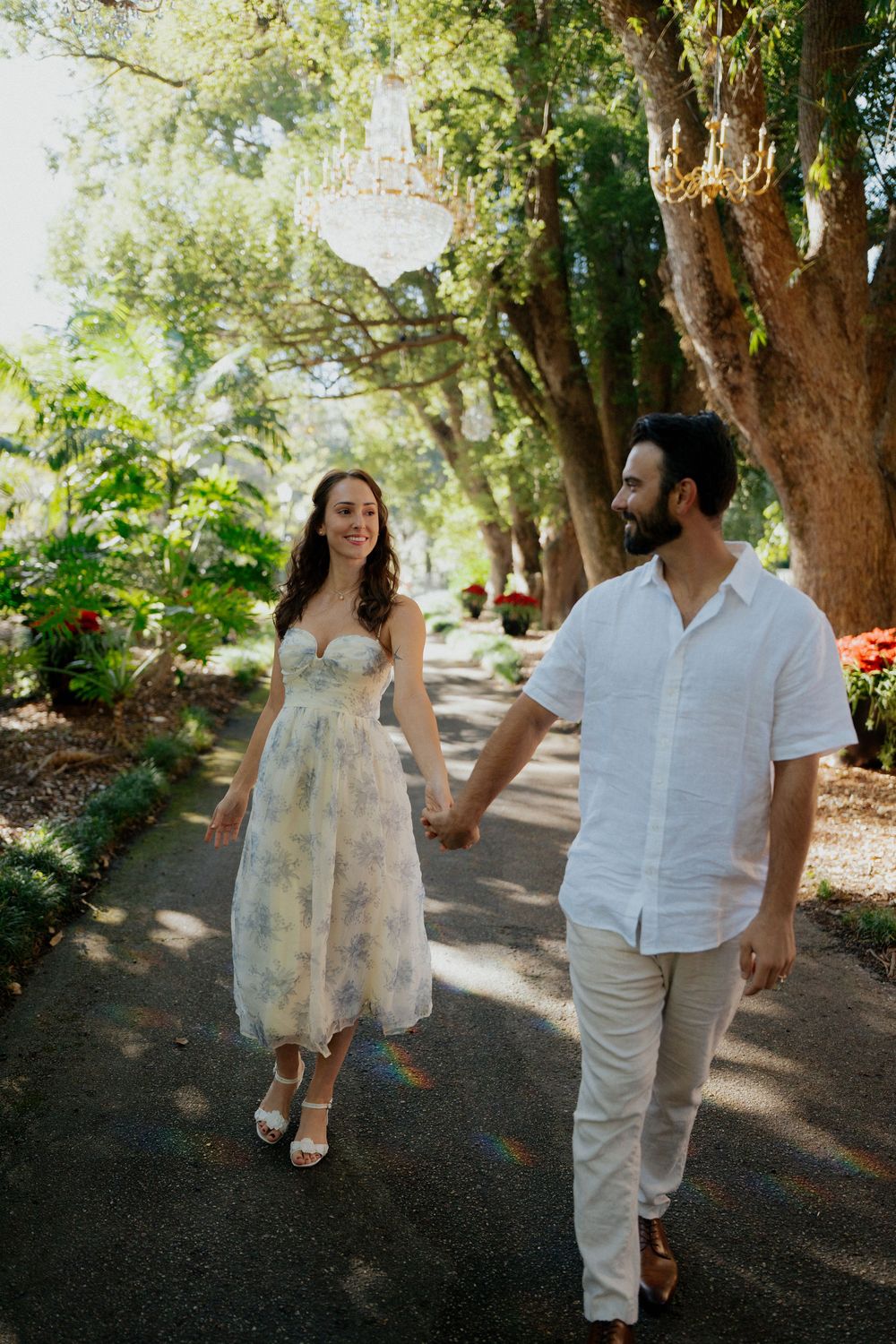 A couple walking together on a garden path, with the woman wearing a floral dress and man in white casual attire.