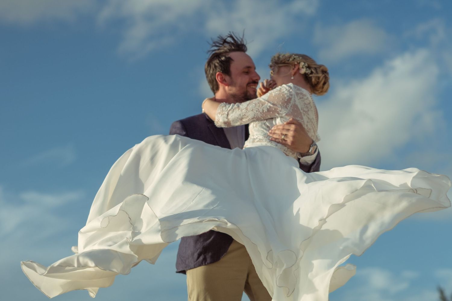 Couple in white outfits dancing on a beach with blue sky and clouds.