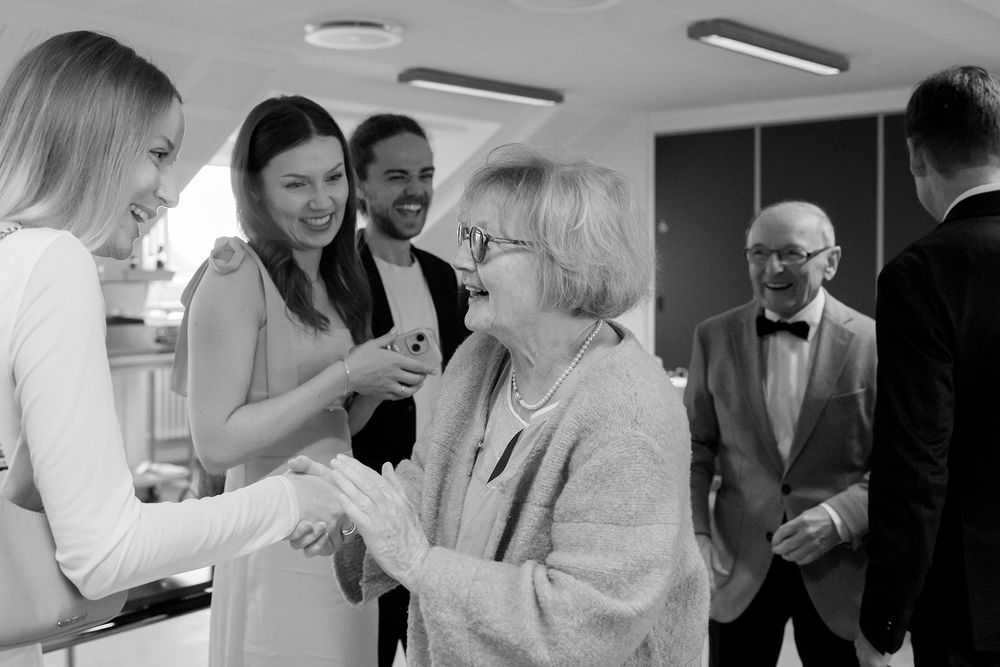 A group of people gather at an indoor event, laughing and socializing in a candid black and white photograph.