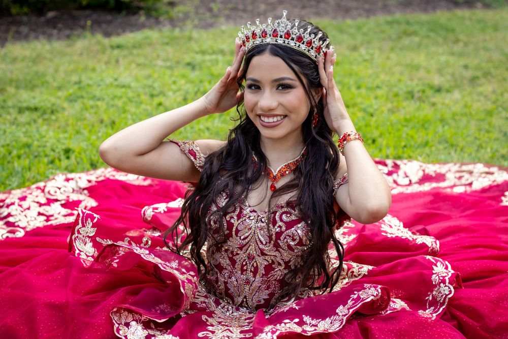 Quinceañera celebration featuring a stunning magenta ball gown and crystal crown on a red blanket outdoors.