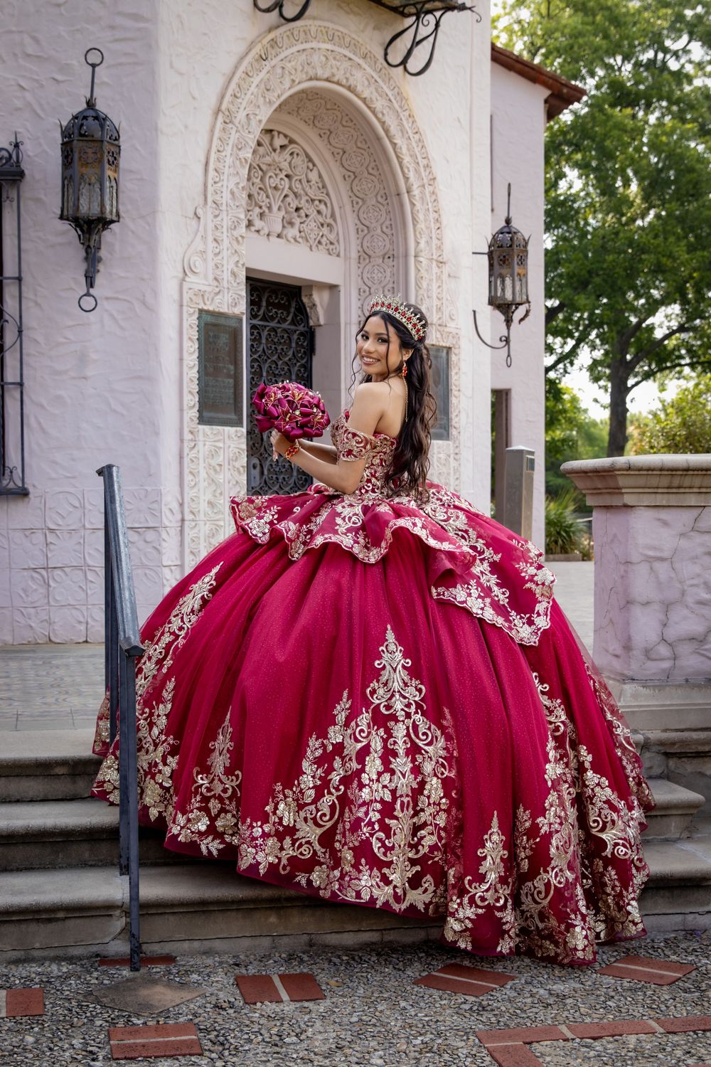 A stunning red quinceañera ball gown with intricate gold embroidery photographed at a Spanish colonial style venue.